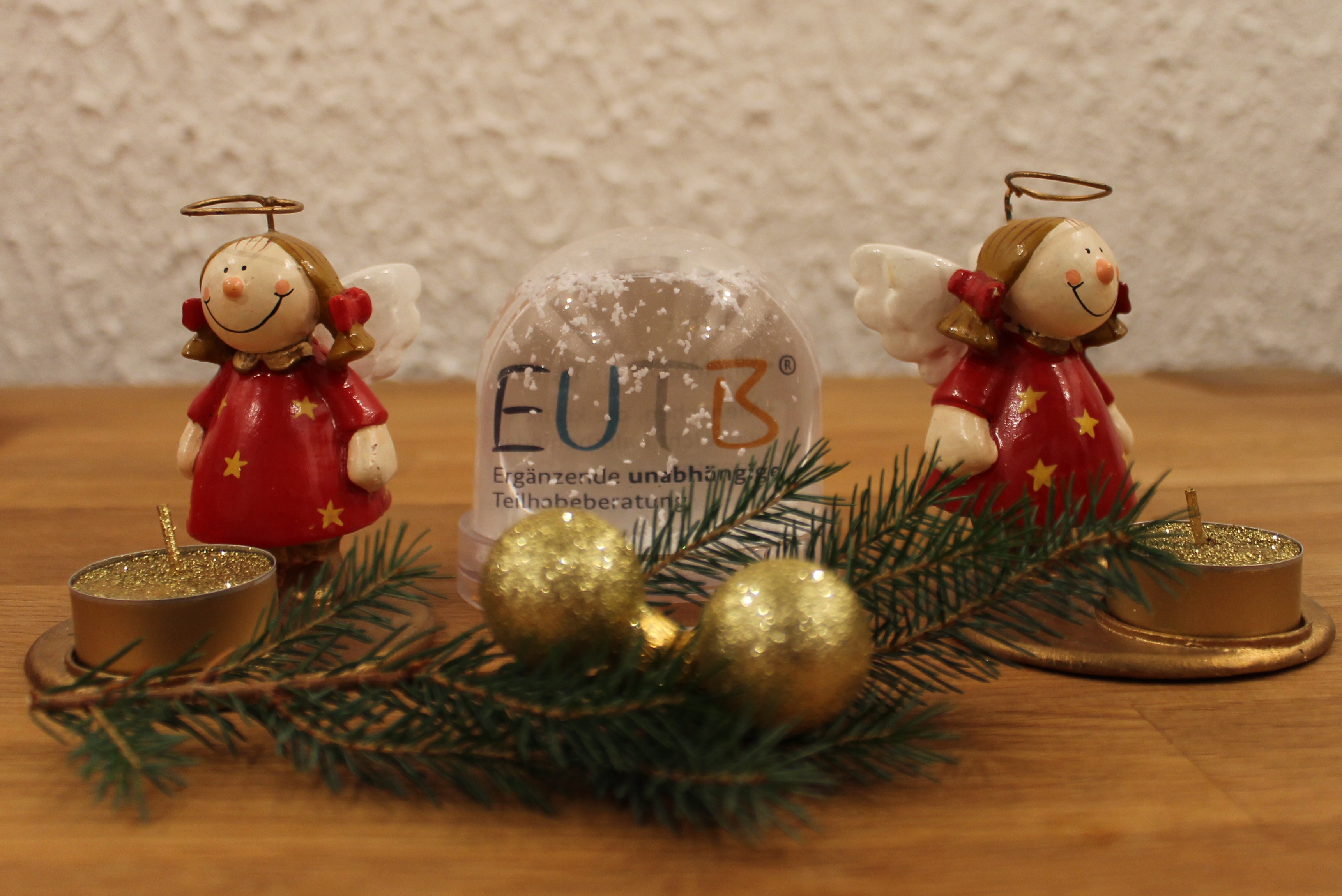 Advents-Dekoration: Zwei Engelfiguren und in der Mitte eine Schneekugel mit EUTB-Logo, davor liegen Zweige, goldene Kugeln und goldene Teelichter.