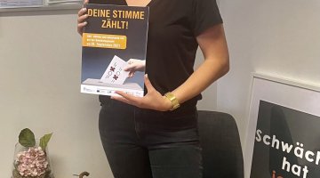 Die Mitarbeiterin der EUTB Paderborn, Annabelle Korn, steht in dem EUTB-Angebot. Sie hält eine Mappe in den Händen. Darauf steht Deine Stimme zählt. Geh‘ wählen und entscheide mit bei der Bundestagswahl am 26. September 2021.