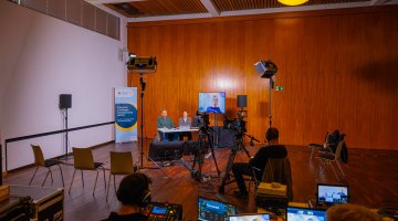 Referierende digital zugeschaltet mit Moderationsduo auf der Bühne. 